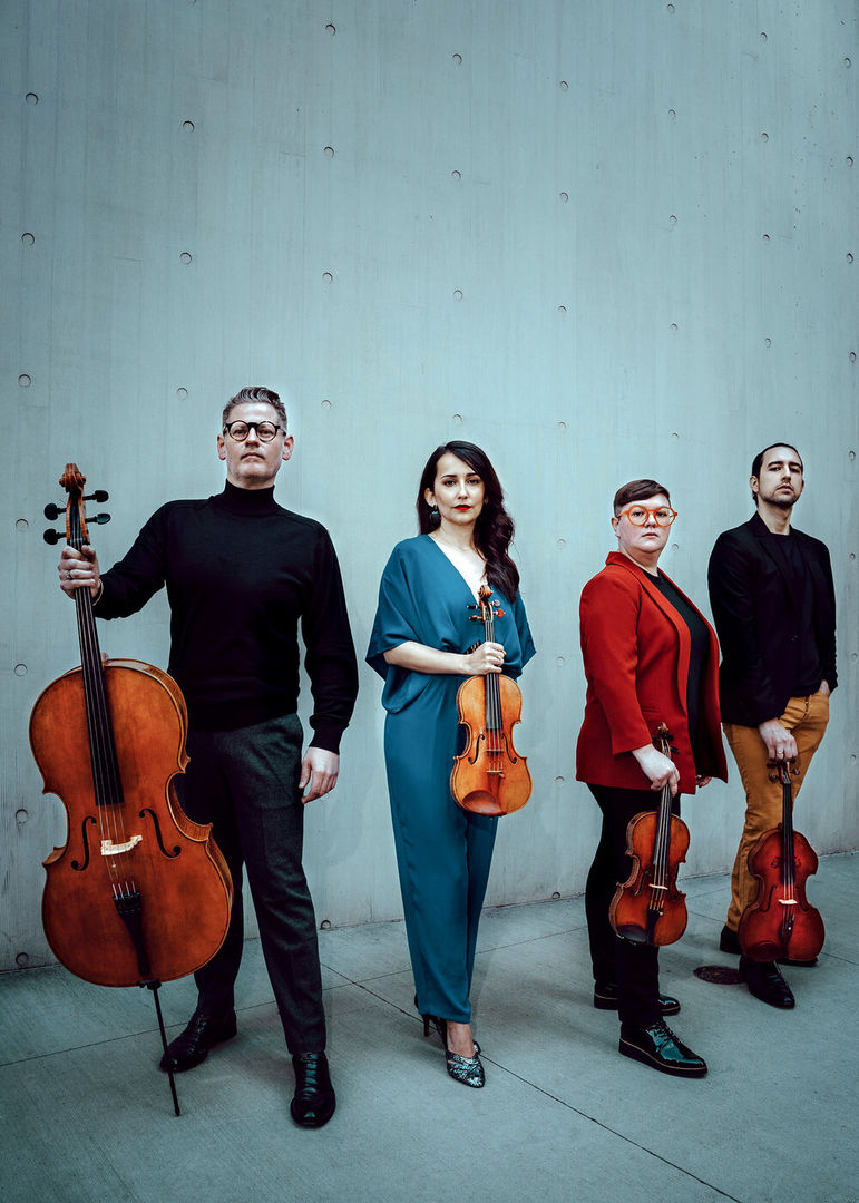 Four members of a string quartet stand in a line against a gray concrete wall. They each hold their instruments and wear chic concert attire in red, blue, and black.